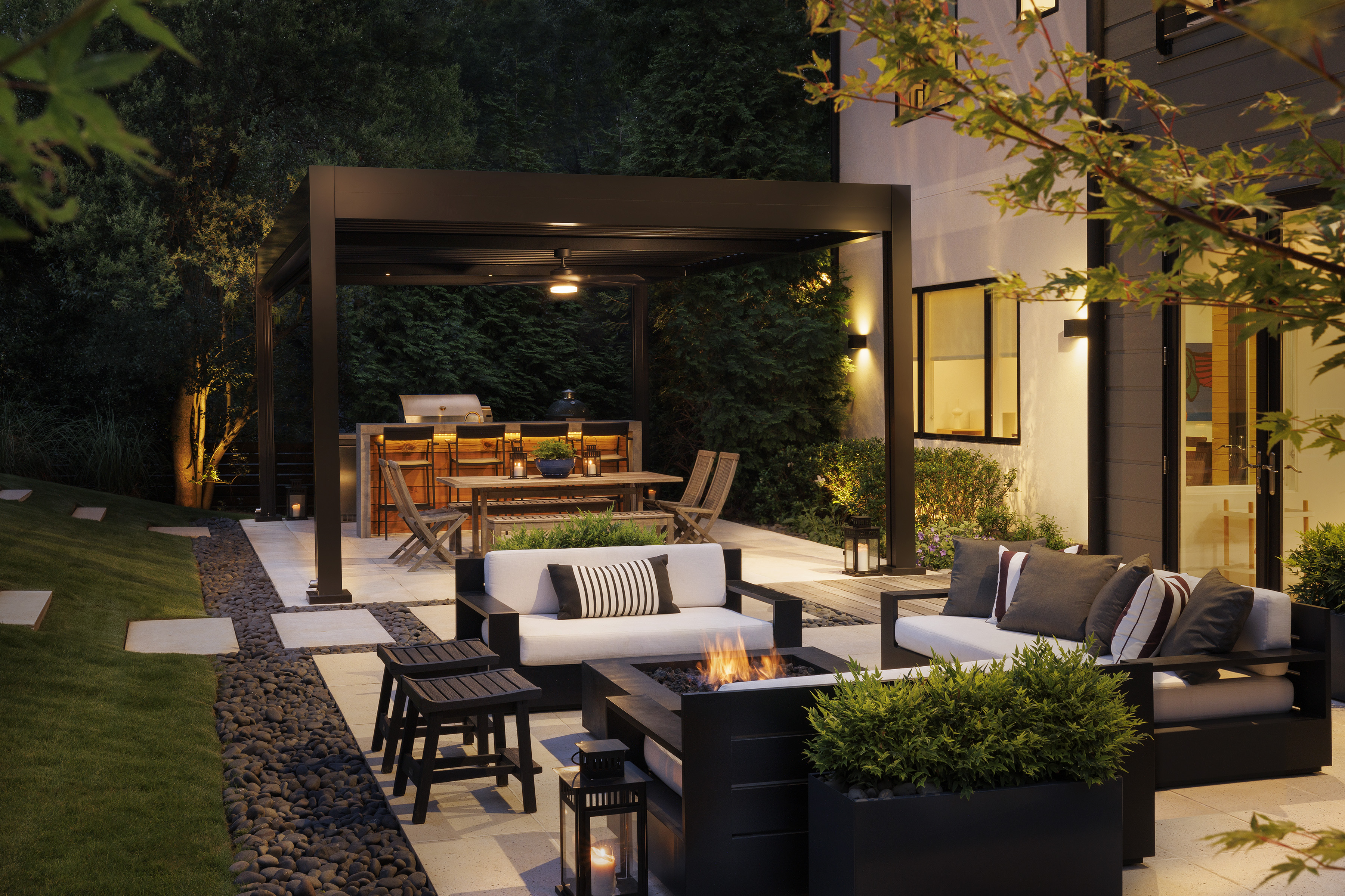 Black R-BLADE pergola at dusk with outdoor kitchen, dining, lounge seating and fire pit